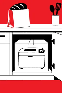 A bulky boxed bread machine wedged into a lower kitchen cabinet, a store-bought loaf in a paper bag on the counter above.